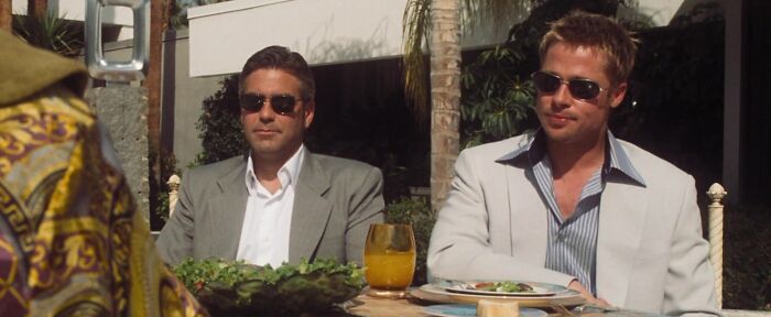 Two iconic best friends in suits and sunglasses, sitting outdoors with a salad and drink on the table.