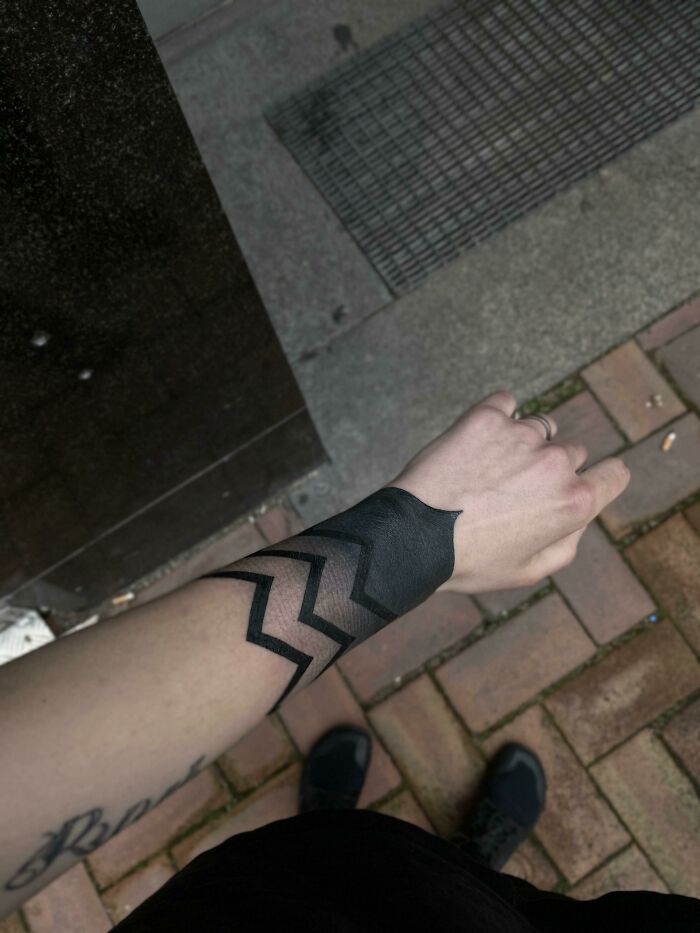 Wrist tattoo with bold black geometric design on a forearm in an urban setting.
