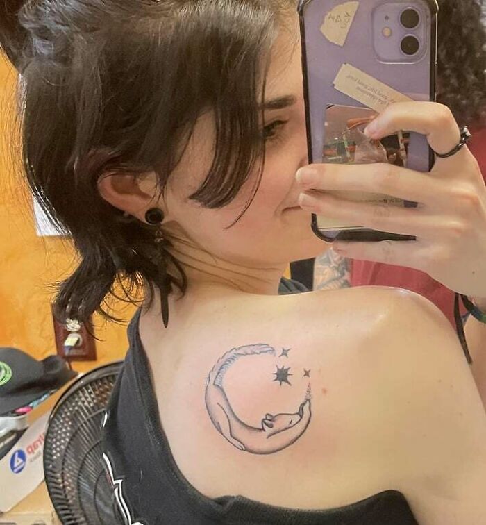 Person showing animal tattoo of a curled cat and stars on their shoulder in a mirror selfie.