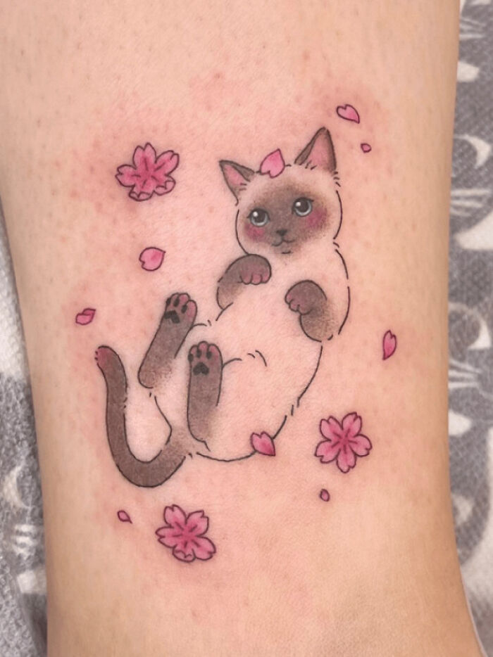 A cute animal tattoo of a playful kitten surrounded by pink flowers and petals on skin.