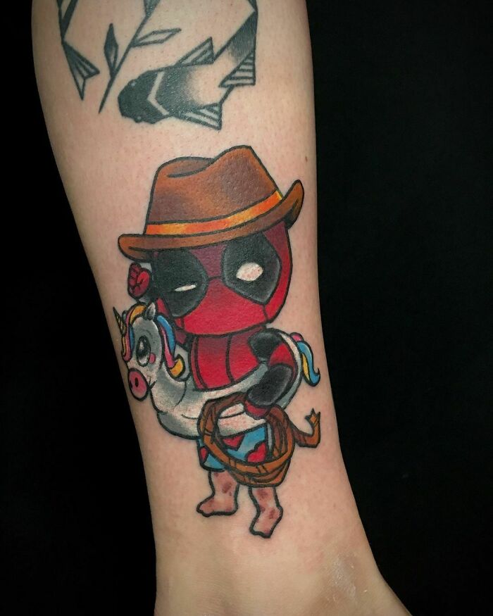Deadpool tattoo wearing a cowboy hat, holding a stuffed unicorn, showcasing vibrant Marvel art.