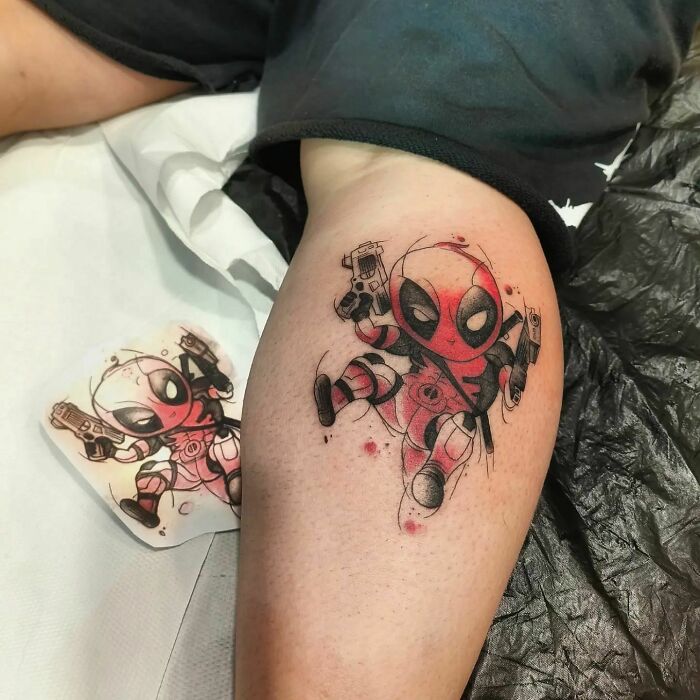 Deadpool Marvel tattoo on leg, featuring vibrant red and black colors, capturing the iconic superhero's playful pose.