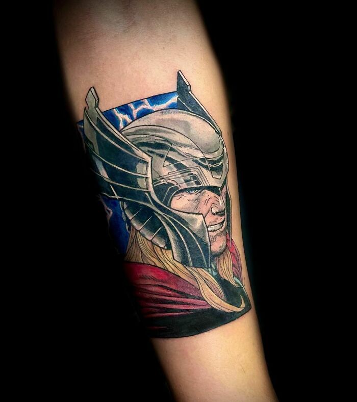 Marvel tattoo of superhero in helmet with flowing blonde hair and red cape on a forearm.