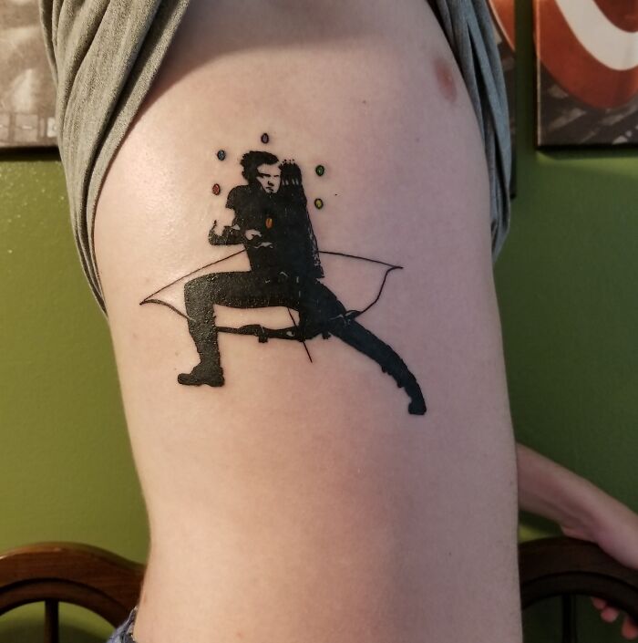 Tattoo depicting a Marvel character Hawkeye with a bow and arrow in a dynamic pose on a person's upper left arm.