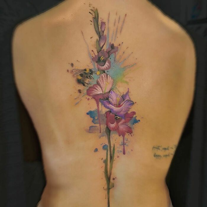 Elegant floral spine tattoo featuring vibrant pink and purple flowers with watercolor effects on a woman's back.