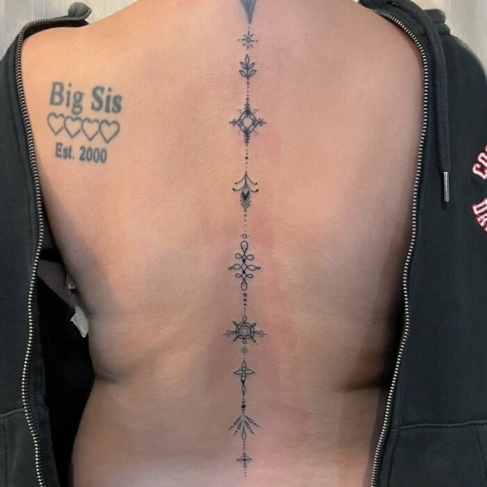 Ornamental spine tattoo with floral motifs, alongside "Big Sis" text and hearts, showcasing intricate body art design.