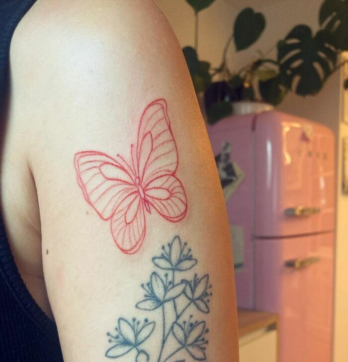 Red ink butterfly tattoo design on arm above a black floral tattoo.