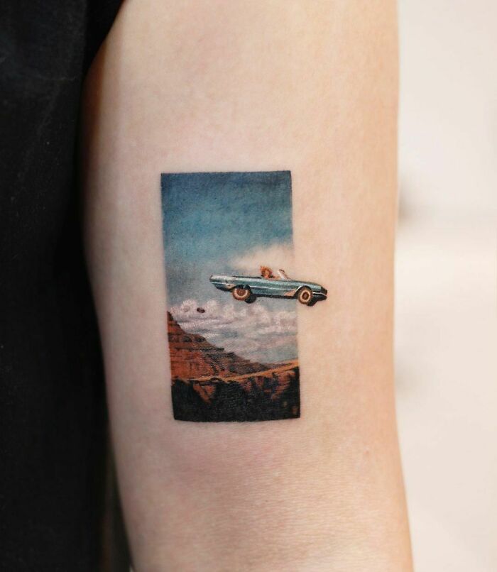 Color tattoo by Saegeem depicting a vintage car soaring over a canyon under a vivid blue sky.