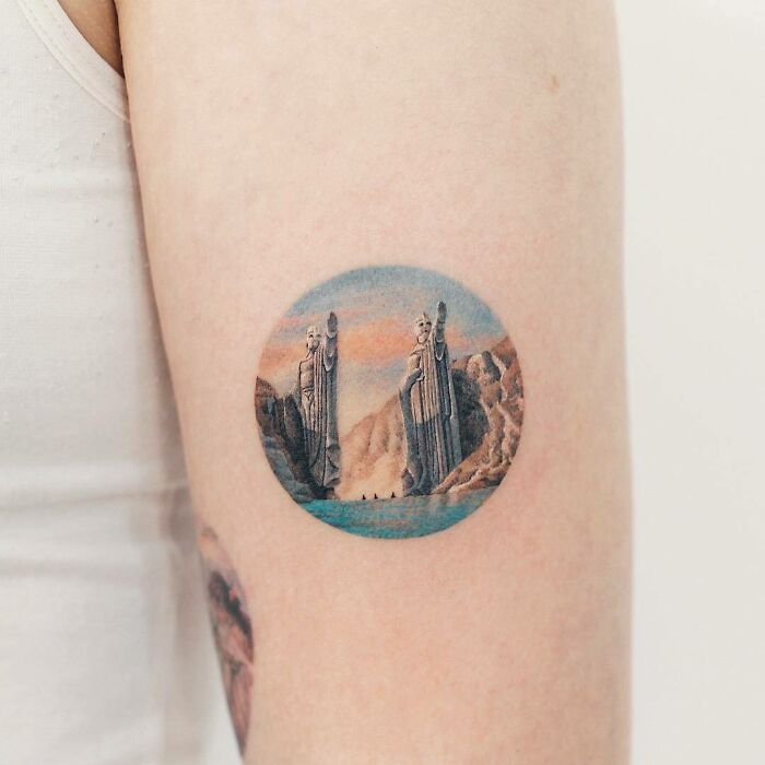 Color tattoo by Saegeem depicting a scenic view with statues and mountains on a person's arm.