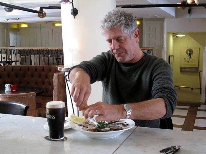 Man squeezing lemon over oysters with a pint of beer on a table, a candid historical photo perspective.