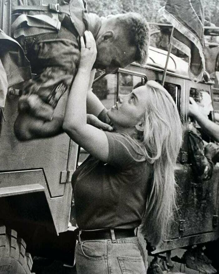 Black and white historical photo of a soldier and woman sharing an emotional moment by a military vehicle with a flag.