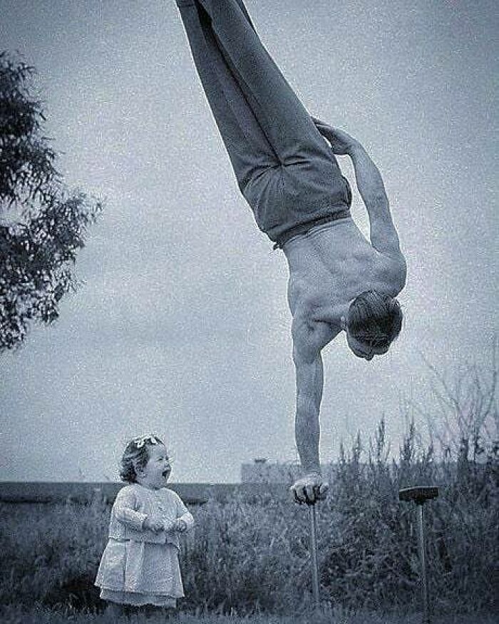 Black and white historical photo of a man performing a one-arm handstand next to a surprised child outdoors.