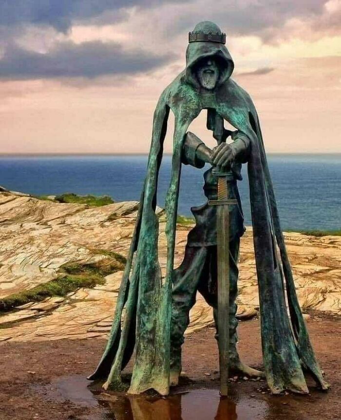 Bronze statue of a hooded medieval knight holding a sword, standing on a rocky coastline under a cloudy sky.