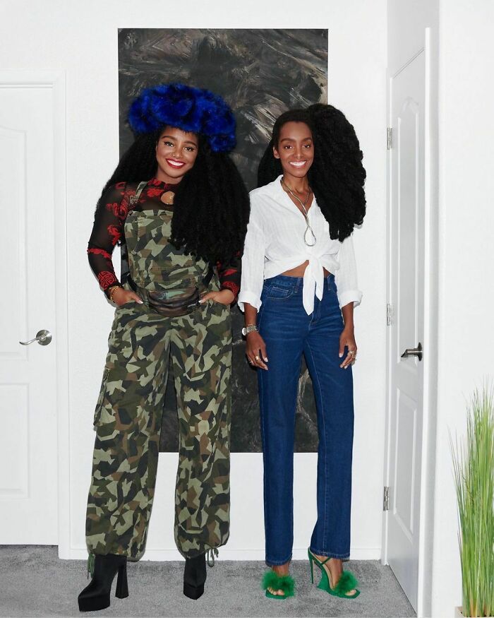 Two stylish celebrity twins with voluminous hair posing together indoors, showcasing unique fashion and confident smiles.