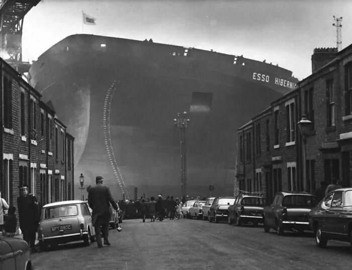 Massive ship towering over narrow street with vintage cars and pedestrians, an important historical photo showing scale contrast.