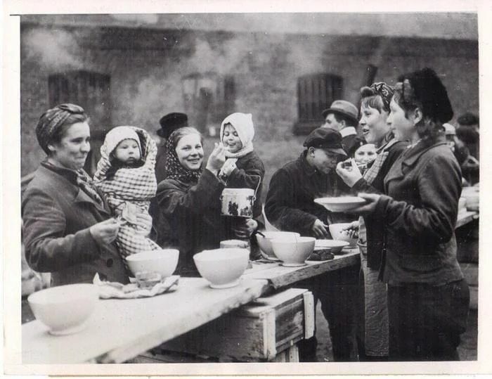 Black and white historical photo showing people, including women and children, gathered around a food line during wartime relief efforts.