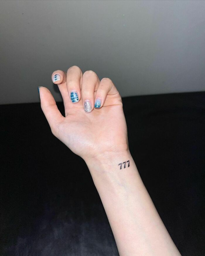 Wrist tattoo of the number 777 on a hand with blue patterned nails.