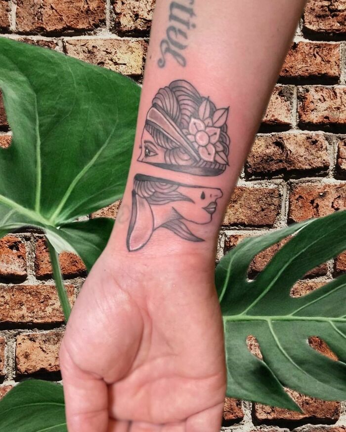 Wrist tattoo featuring a woman's face design with floral elements, set against a leafy and brick background.