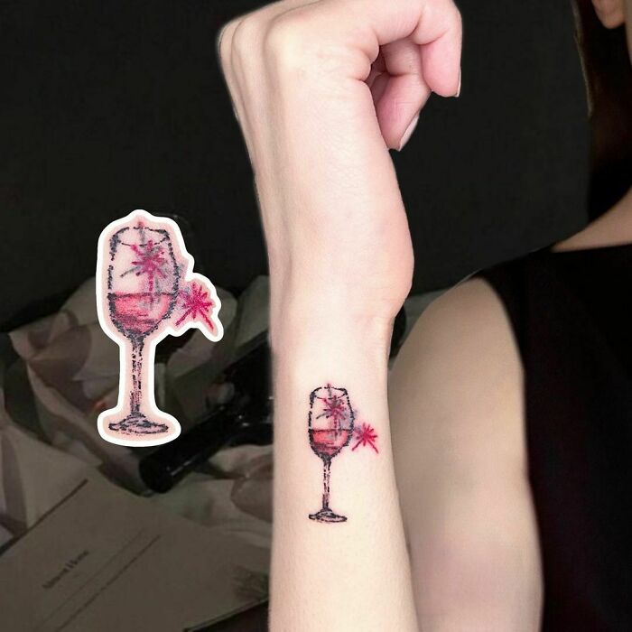 Wrist tattoo of a glass with red liquid and flower design.
