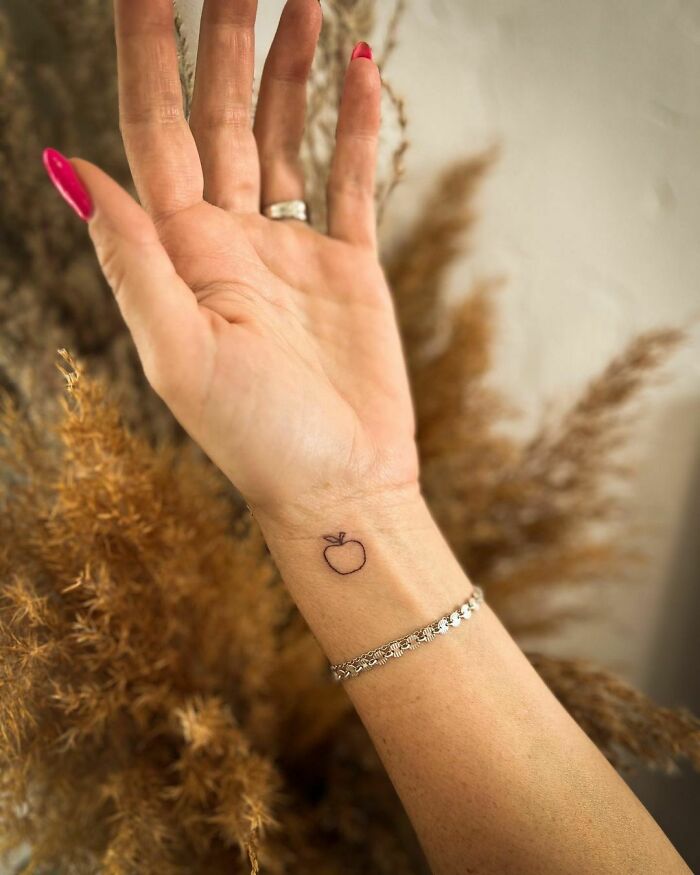 Small apple wrist tattoo with a silver bracelet, against a dried flower backdrop.