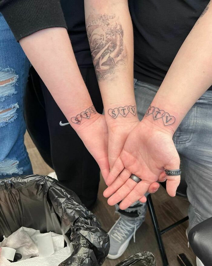 Two people with wrist tattoos featuring intricate designs and letters inside heart shapes.