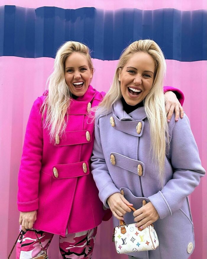 Two smiling celebrity twins wearing bright coats posing against a pink and purple backdrop outdoors.