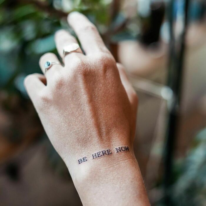 Minimalist wrist tattoo with text "Be Here Now," exemplifying statement-making design.