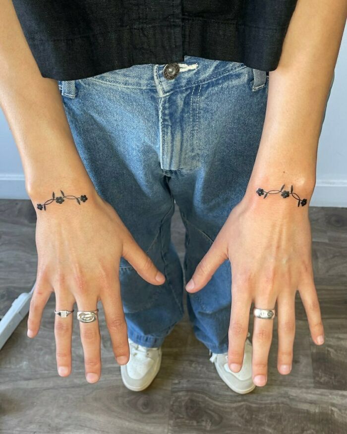 Person with floral wrist tattoos and rings, wearing jeans and sneakers.