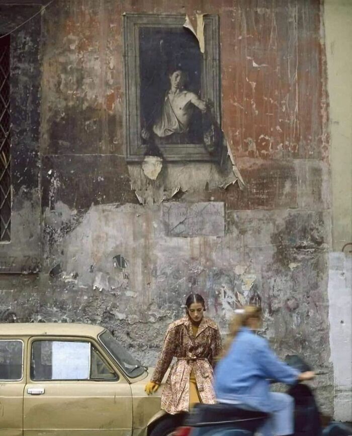 A peeling mural of a classical portrait above a woman by a vintage car, with a blurred figure on a scooter passing by.