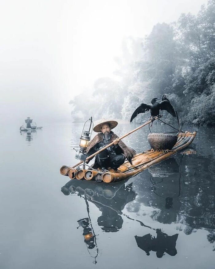 Man on bamboo raft with cormorant birds, wearing a traditional hat, on misty river; amusing Instagram photo.