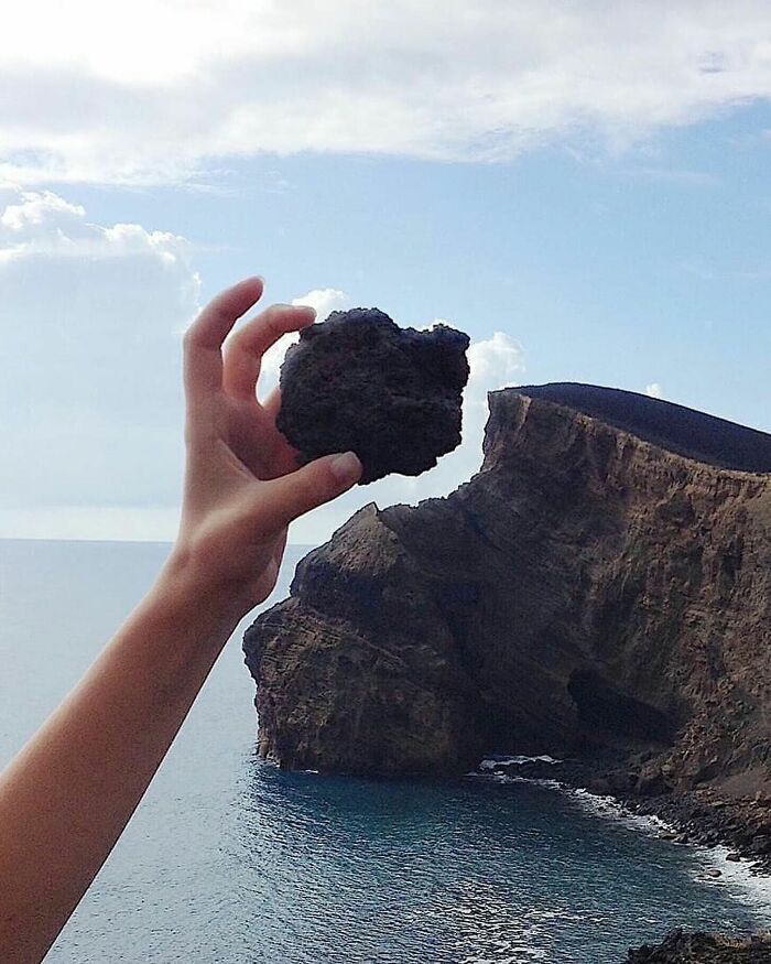 Hand holding a rock that matches the shape of a cliff in the background, highlighting amusing photo creativity.