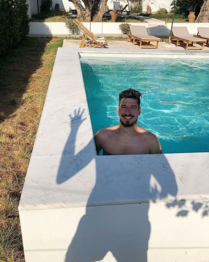 Man in pool waves as shadow falls on the ground, creating an amusing photo moment.