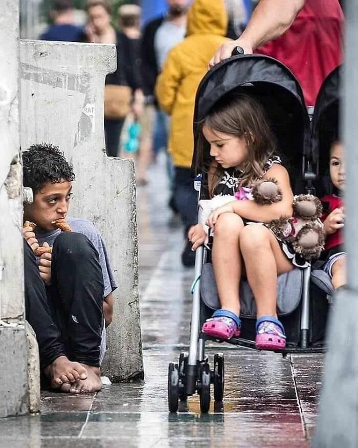 A boy sitting barefoot eating bread beside a child in a stroller, capturing a moment of contrast on a busy street.