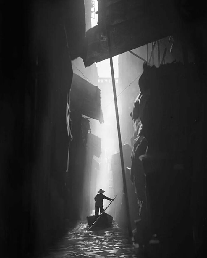 Person rowing a boat through a narrow, shadowy alley, illuminated by soft light. Instagram page photography highlights.