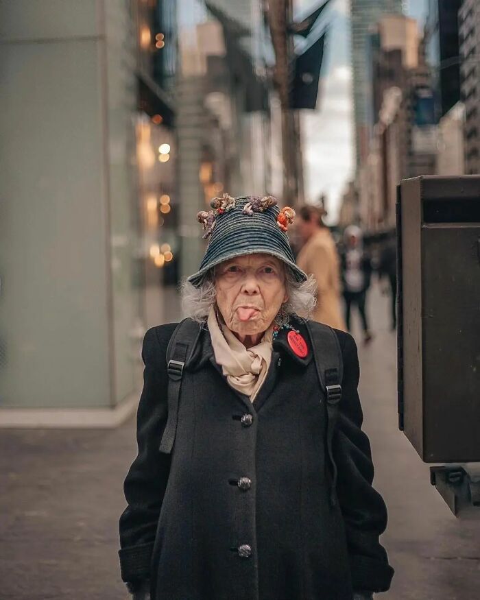 Elderly woman in a city street, wearing a hat adorned with flowers, making a playful face. Amusing Instagram photo.