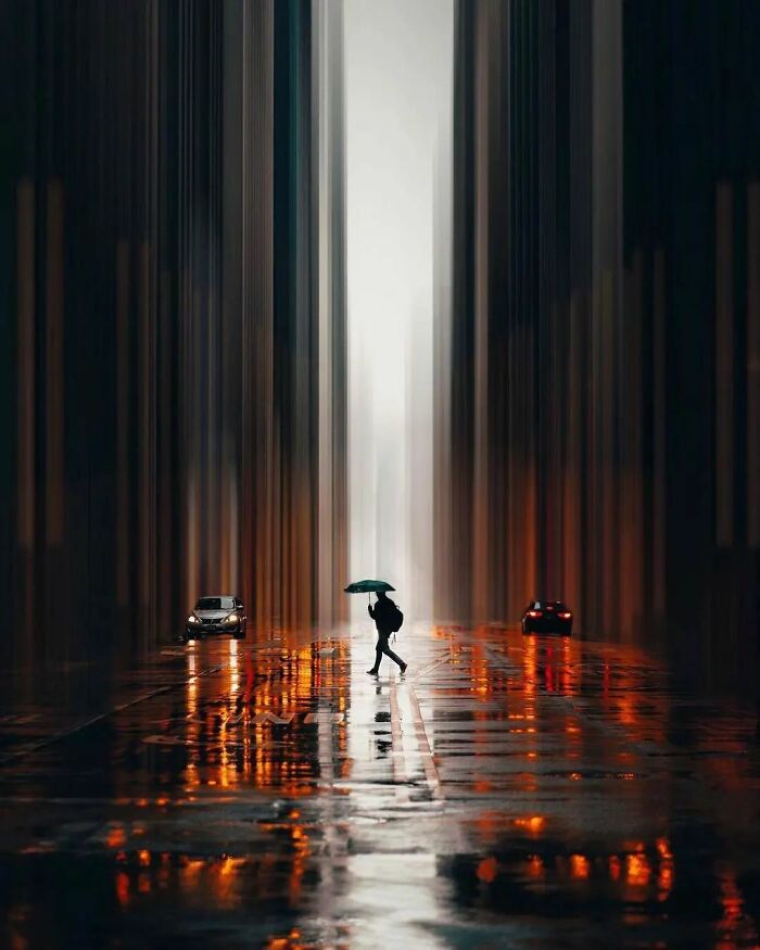Umbrella holder walking between cars on rainy street in artistic cityscape, reflecting colors, part of amusing photo collection.