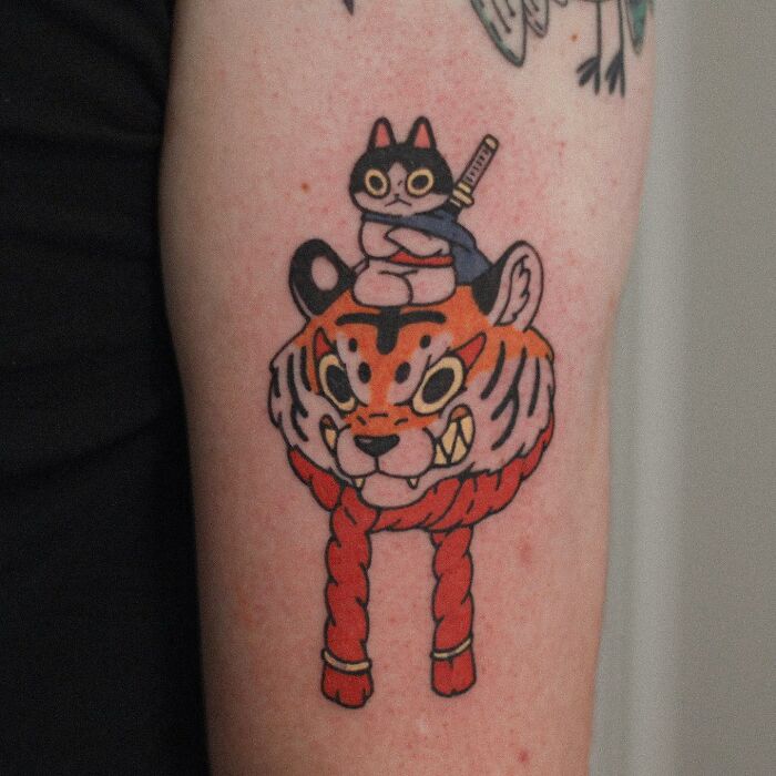 Tattoo of a cute cat with a sword sitting atop a tiger's head, showcasing animal-themed design.