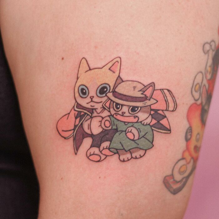Adorable animal-themed tattoo featuring two cartoon cats dressed in cute outfits, sitting together.