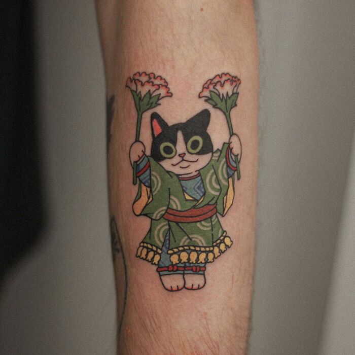 Tattoo of a cute cat in traditional attire holding flowers, showcasing an adorable animal-themed design.