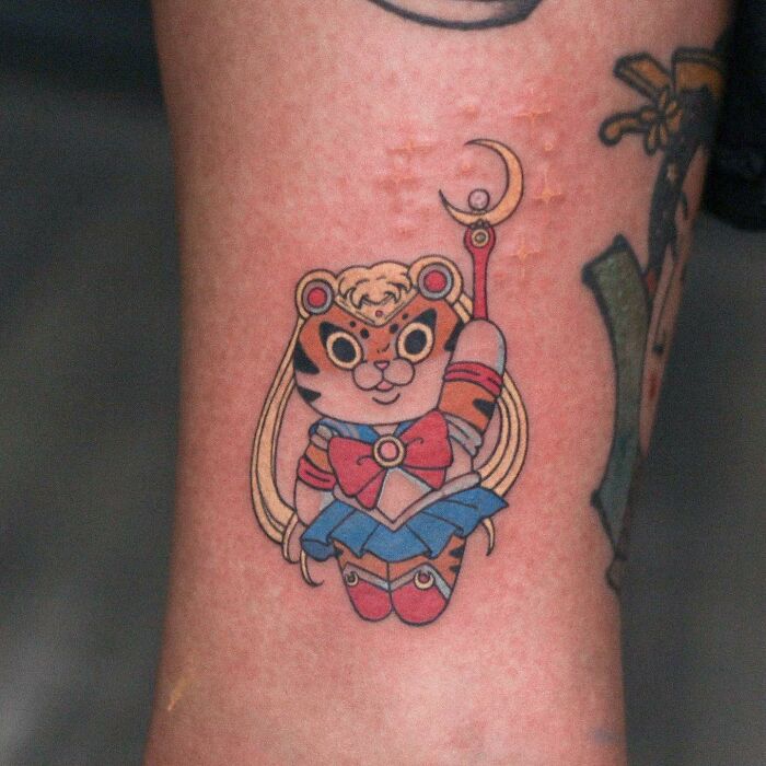 Adorable animal-themed tattoo of a cartoon-style tiger with long hair and sailor outfit on skin.