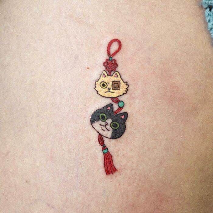 Animal-themed tattoo featuring two cute cat faces, one yellow and one gray, connected by a decorative red tassel.