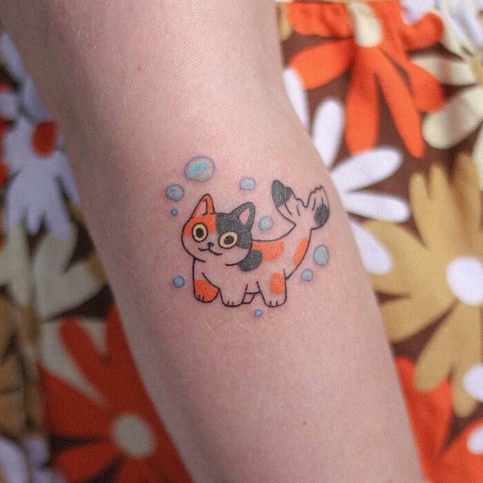 Adorable animal-themed tattoo featuring a colorful cat-mermaid design on a person's arm.