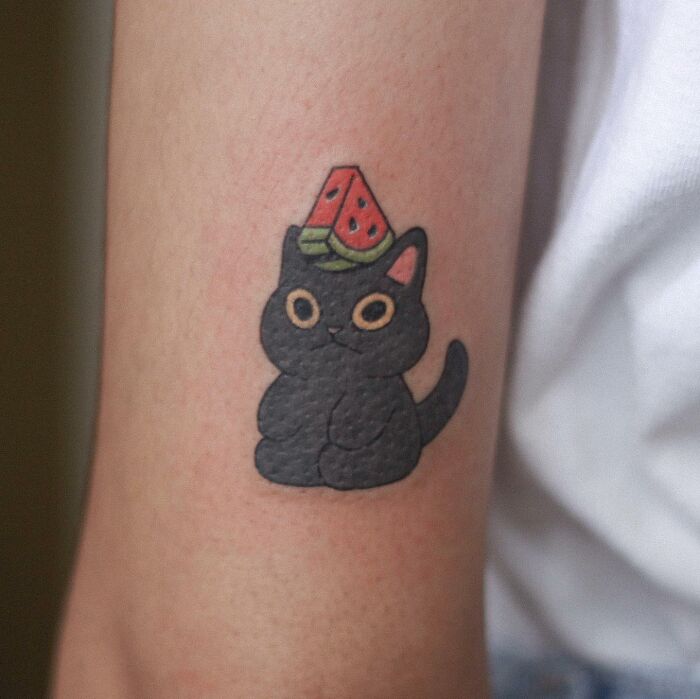 Adorable animal-themed tattoo of a black cat with a watermelon hat on skin.