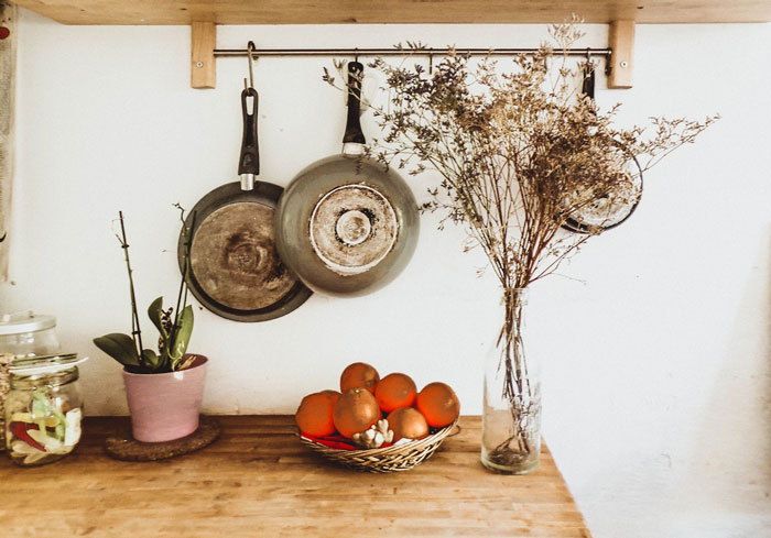 Home decoration with hanging pans, vase of dried flowers, and basket of oranges on a wooden countertop.