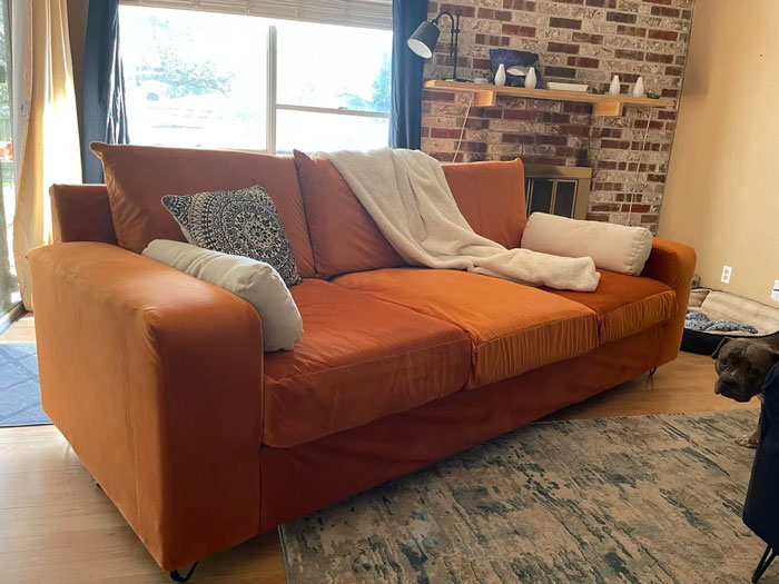 Orange sofa with assorted cushions, white throw, near a brick wall in a cozy living room; home decoration idea.
