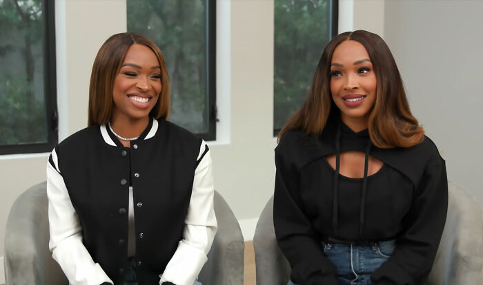 Two celebrity twins sitting indoors, smiling and wearing casual black and white outfits, showcasing celebrity twins.