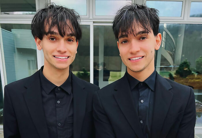 Celebrity twins smiling side by side in matching black outfits standing outdoors near a windowed building.
