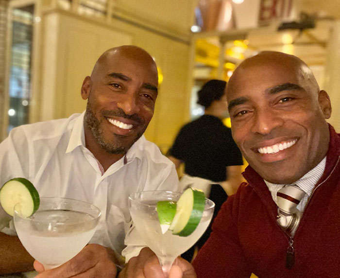 Two celebrity twins smiling and holding cocktails with cucumber garnish in a warmly lit restaurant setting.