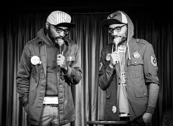 Celebrity twins performing on stage, each holding a microphone and wearing casual streetwear with hats and glasses.