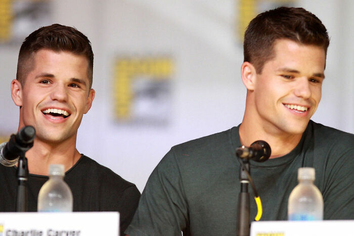 Two smiling celebrity twins speaking into microphones at a panel, showcasing celebrity twins who prove great things come in pairs.